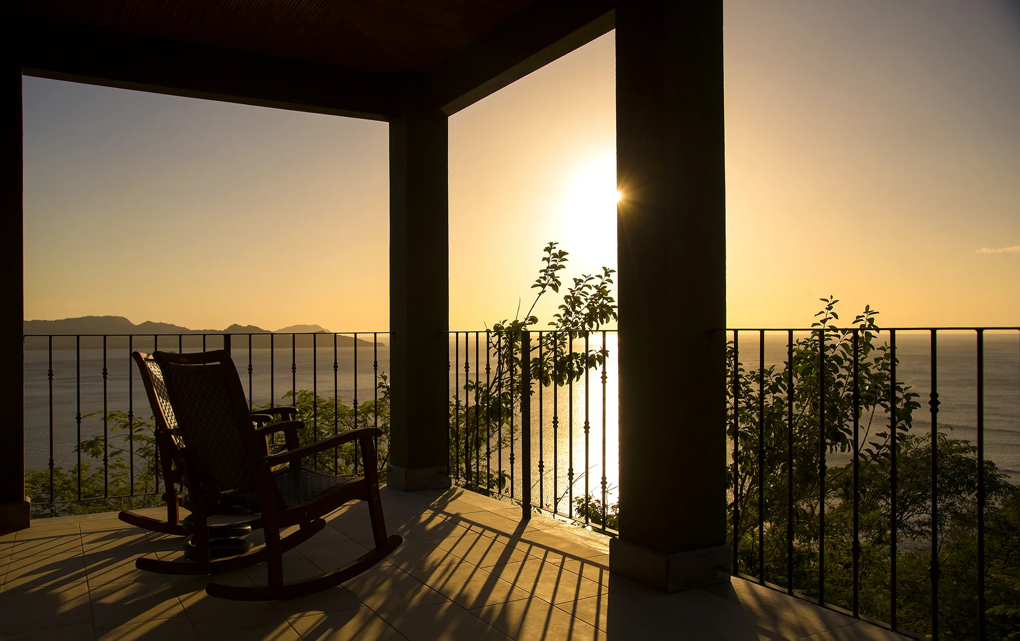 A rocking chair on a 360 Splendor terrace overlooking the Pacific at sunset, with the headland and islands of Playa Flamingo in the distance.