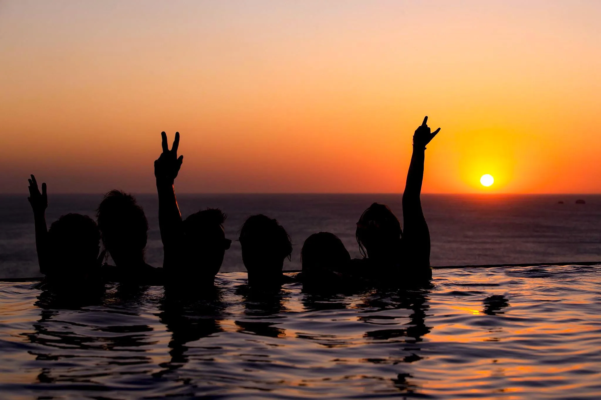 A group of friends in the infinity pool at 360 Splendor raise their arms in celebration as the sun sets over the Pacific.